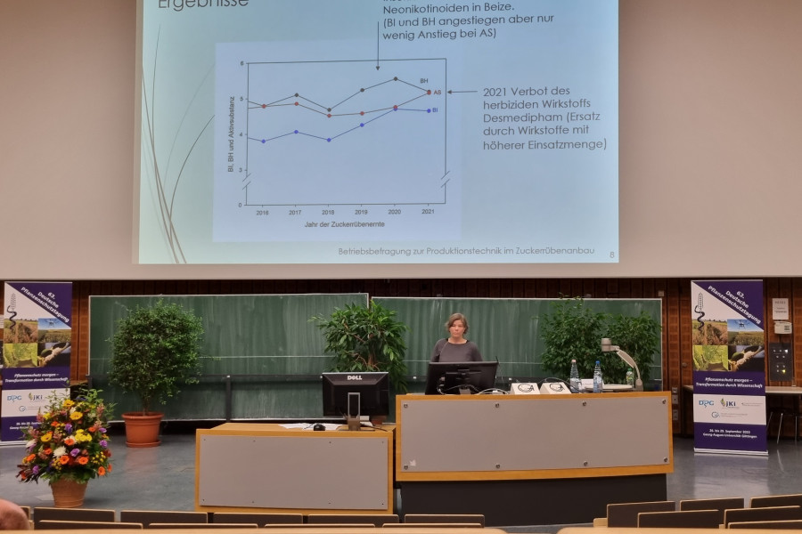 Vortrag einer Frau am Rednerpult auf einer Tagung, PowerPointfolie im Hintergrund mit Kurvendiagramm