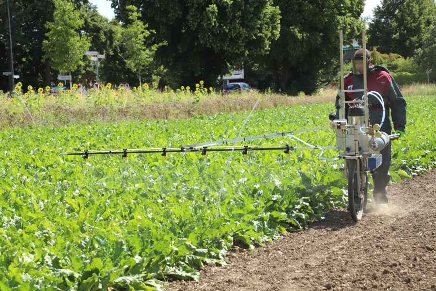 Mann im Vollschutzanzug schiebt eine Fahrradspritze: ein Einrad mit Halterung und Spritzbalken an der Seite