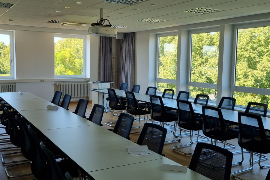 ein Sitzungsraum mit weißen Tischen, Stühlen, Fensterreihe mit grünen Bäumen